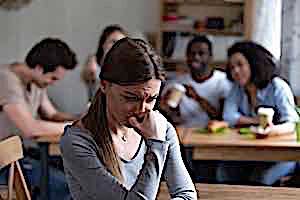 sad scared woman sitting alone with group of friends sitting at different table