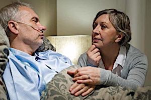 sad older woman sits by hospital bed of elderly man