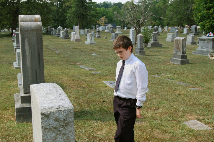 sad boy in cemetary looking at grave stone