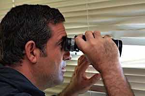 man with binoculars spying through window blinds