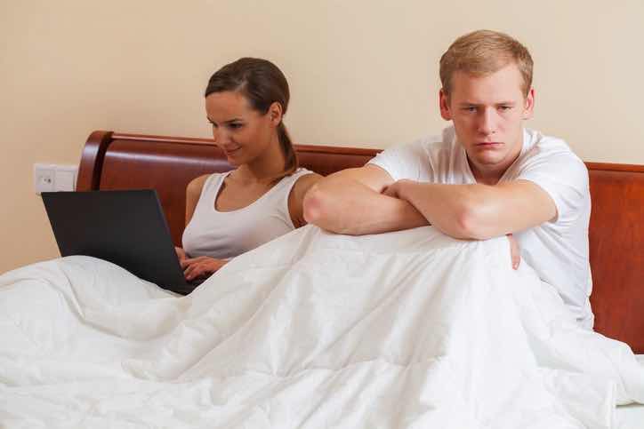 angry man in bed with back to woman on computer no sex