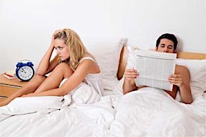 man reads paper in bed next to frustrated woman 