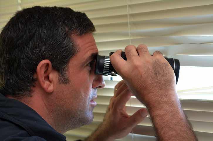 suspicious man peering through blinds with binoculars