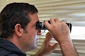 suspicious man peering through blinds with binoculars